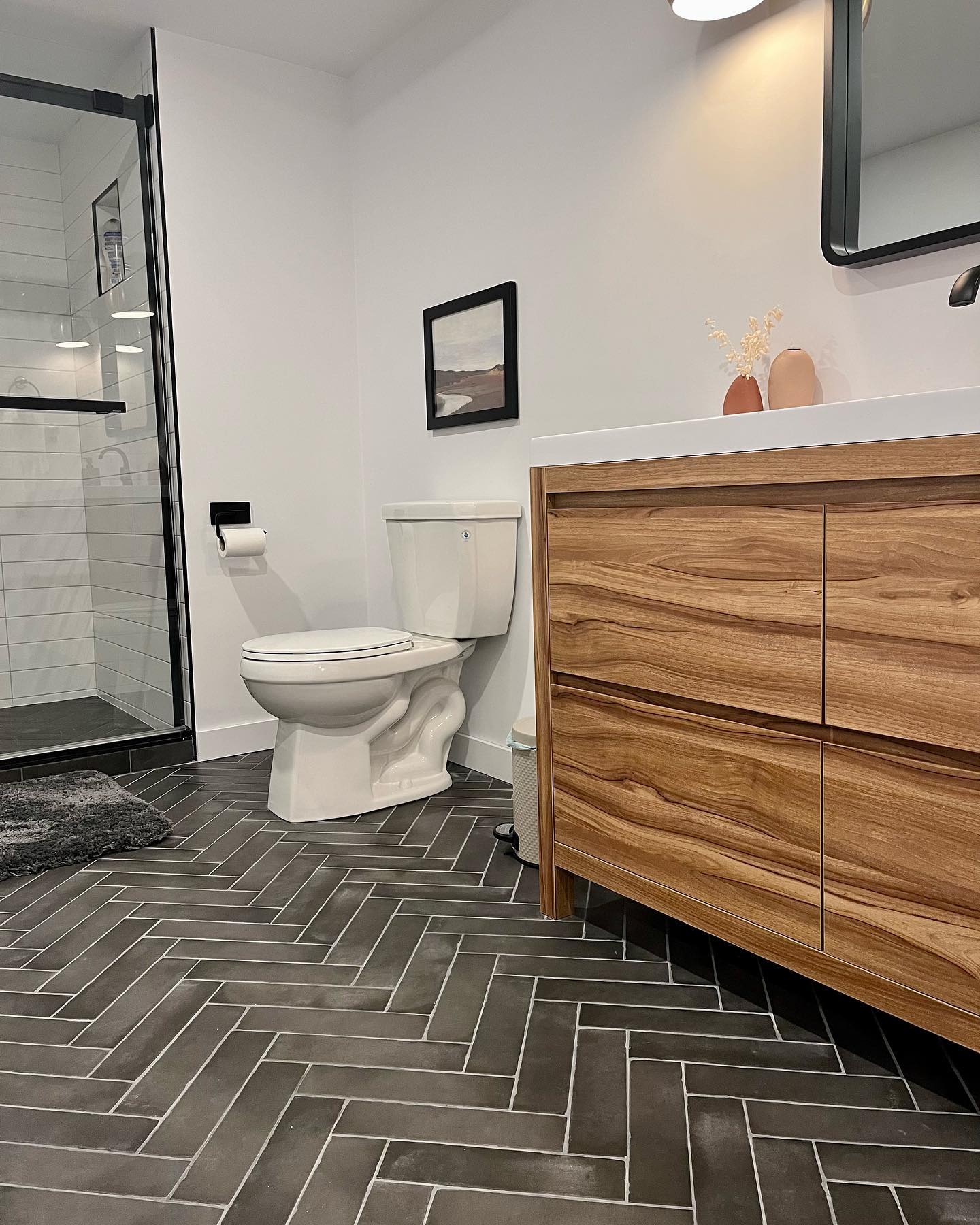 Wood vanity with dark gray herringbone floor and black framed shower enclosure in new basement bathroom
