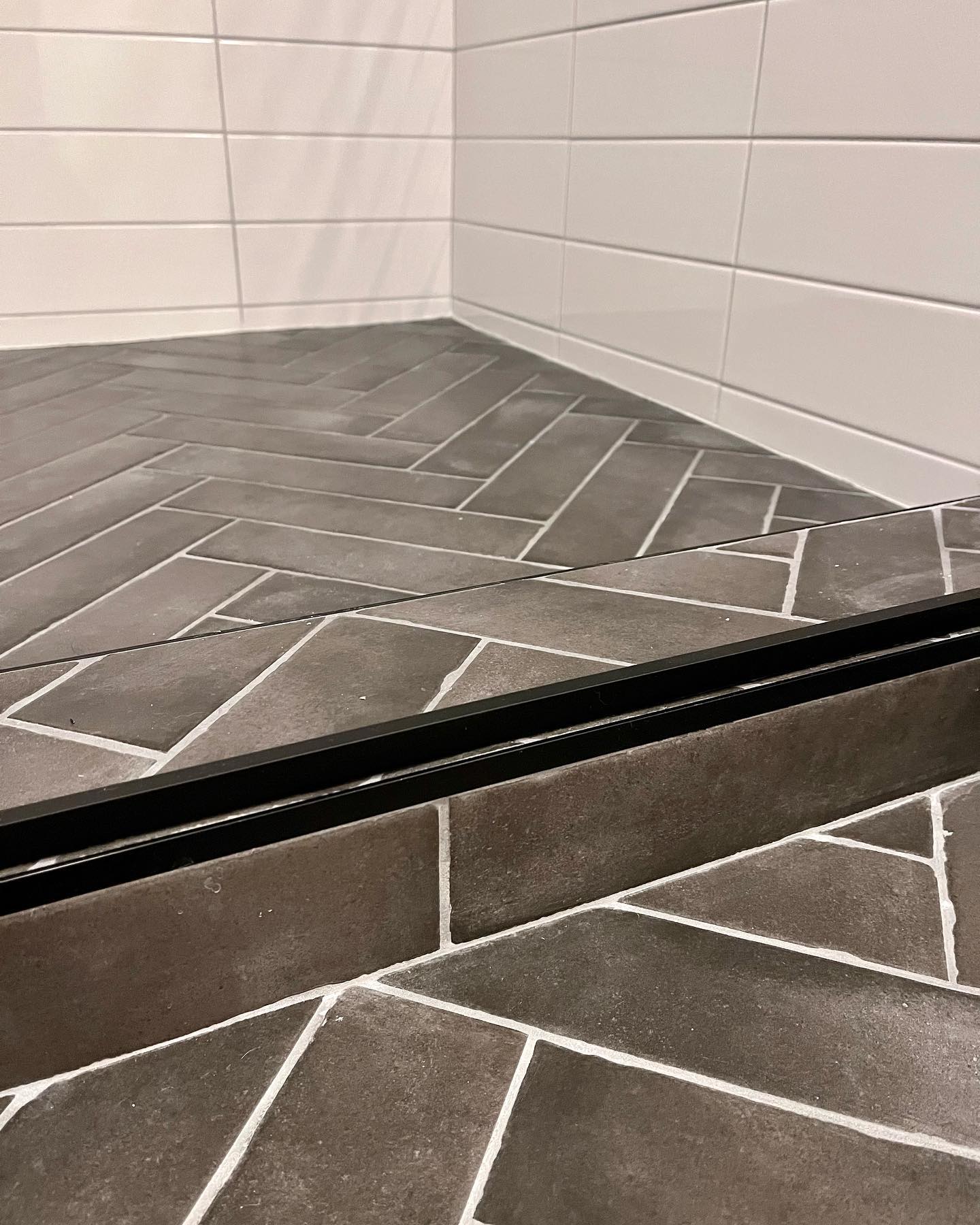 Black framed sliding glass shower enclosure with white subway tile surround over dark gray herringbone floor